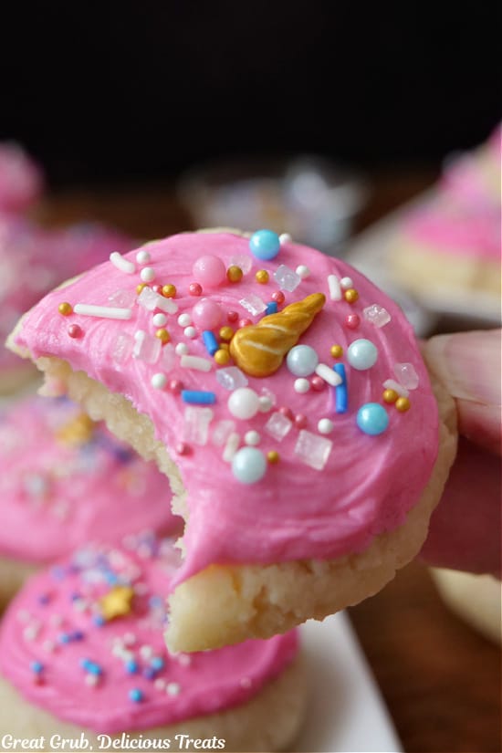 A close up of a sugar cookie with pink frosting and candy sprinkles that has a bite taken out of it.