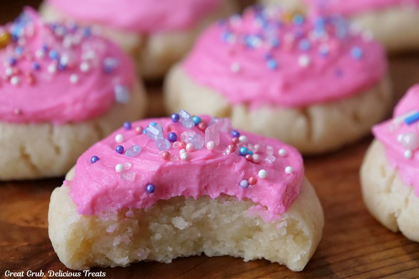 A close up of a wood surface with a few sugar cookies with pink frosting and candy sprinkles on top with a bite taken out of the cookie in front.