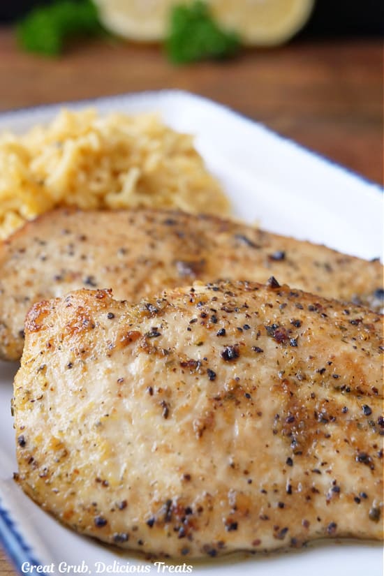 A close up of two boneless skinless lemon pepper chicken breasts on a white plate with a side of rice.