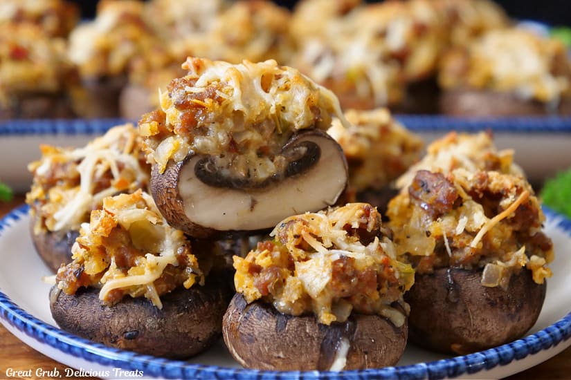 A horizontal photo of a round white plate with blue trim filled with a half dozen stuffed mushrooms with more in the background.