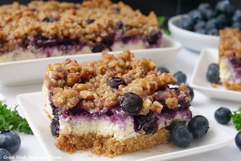A horizontal photo of blueberry cheesecake bars with one on a white plate with loose blueberries on the plate.