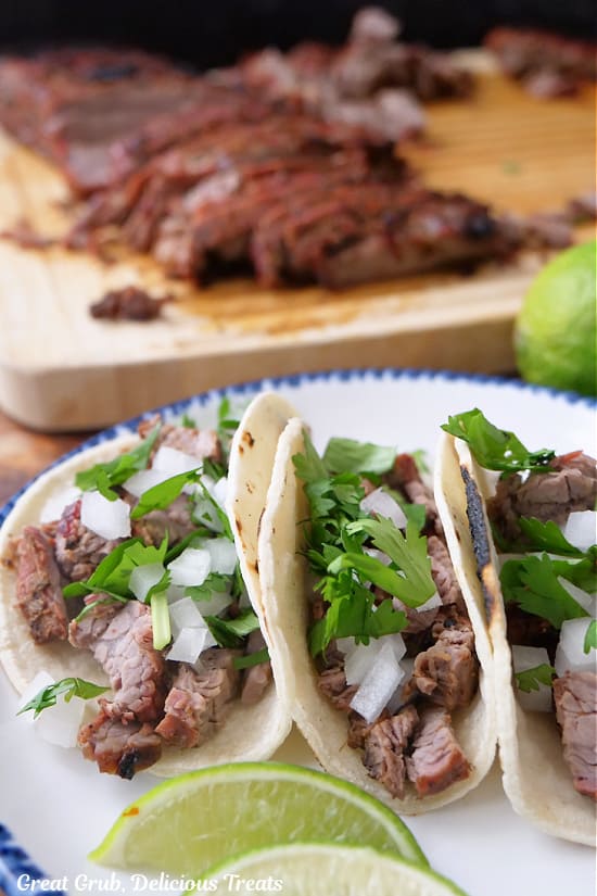 Three street tacos on a white plate with blue trim with carne asada on a cutting board in the background.