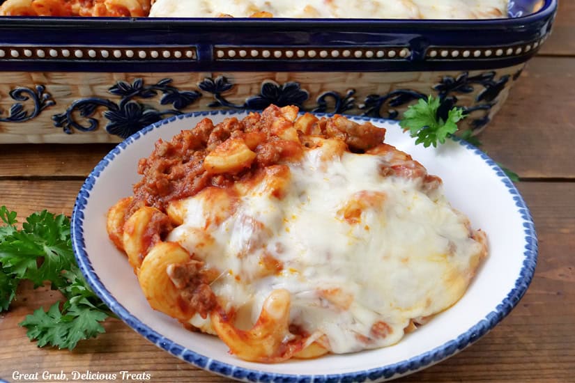 A horizontal photo of a wood surface with a white bowl with blue trim filled with a serving of pasta with melted cheese and a meat sauce.