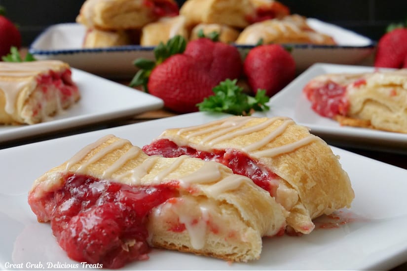A horizontal photo of strawberry cream cheese pastry slices on white plates and fresh strawberries in the background.