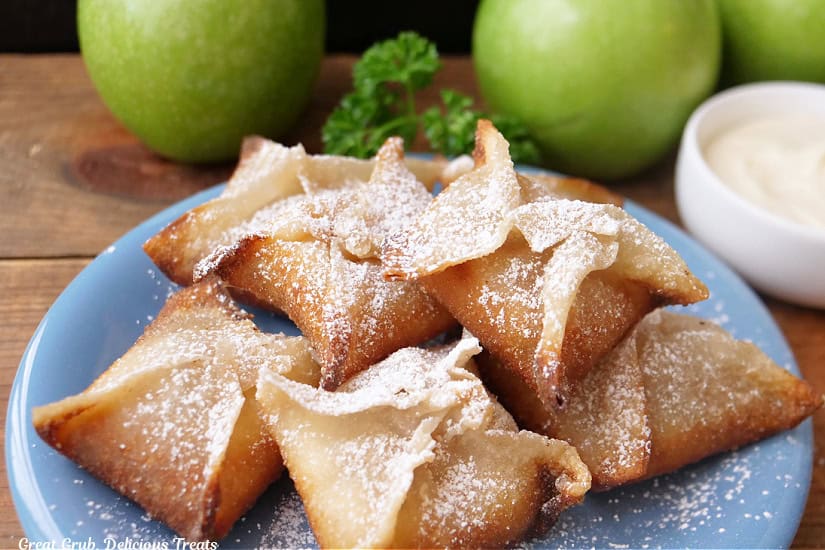 A light blue plate with seven apple pie wontons on it with green apples in the background.