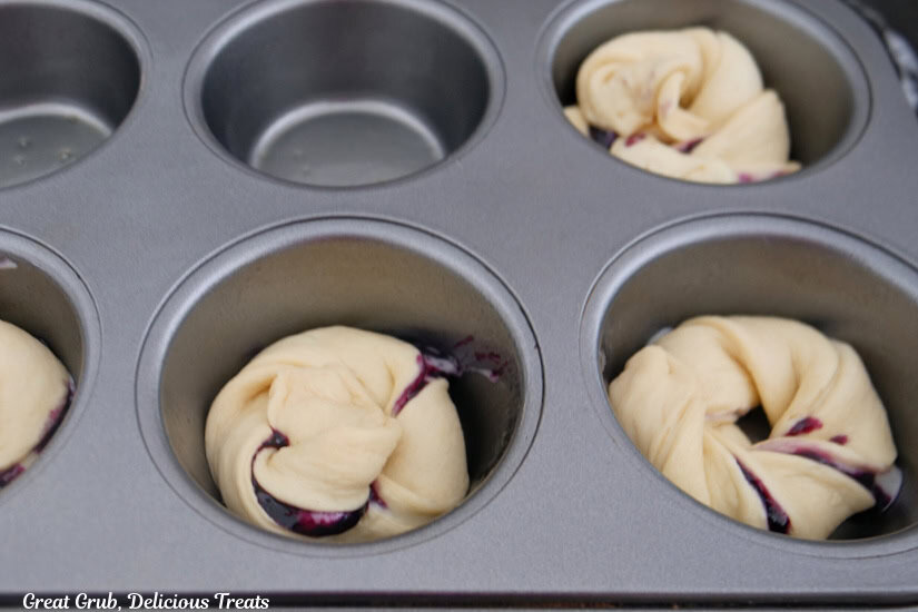 A silver muffin tin with twisted crescent dough filled with cream cheese and blueberry filling placed in the tin.