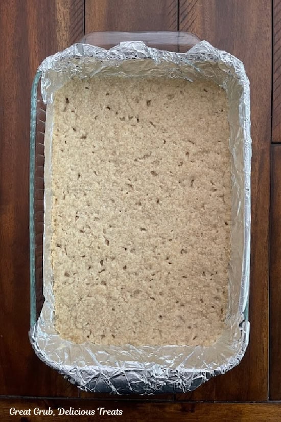 A dark wood surface with a foil lined 9x13 glass baking dish with a shortbread crust in it.