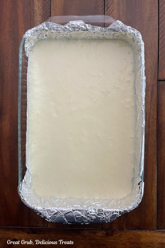 A dark wood surface with a foil lined glass baking dish with a layer of cheesecake pour over a shortbread crust.