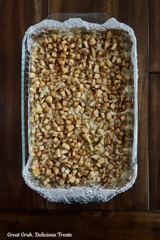 A dark wood surface with a foil lined glass baking dish with chopped apple and cinnamon layered on top of the cheesecake layer.