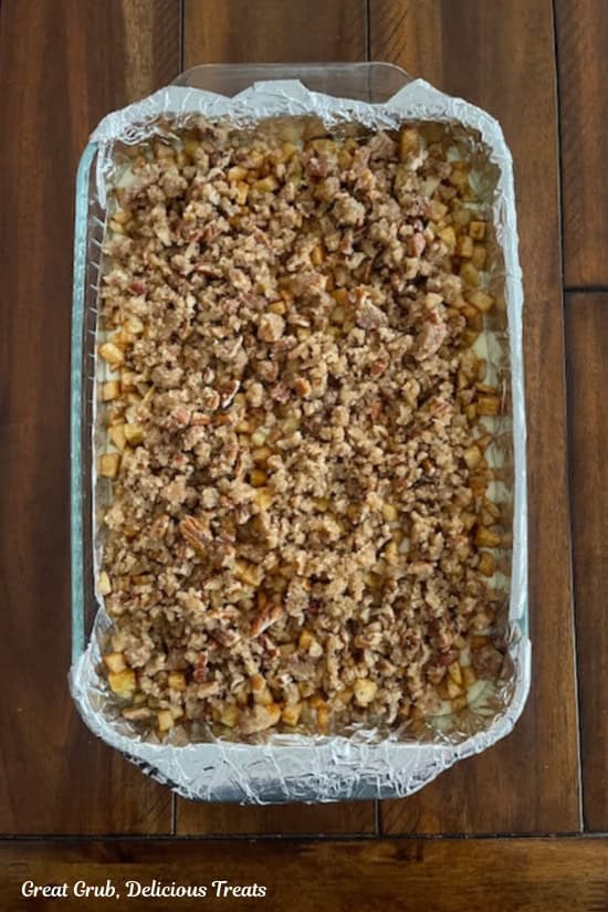 A foil lined glass baking dish on a wood surface with brown sugar pecan topping on top of the chopped apple layer.