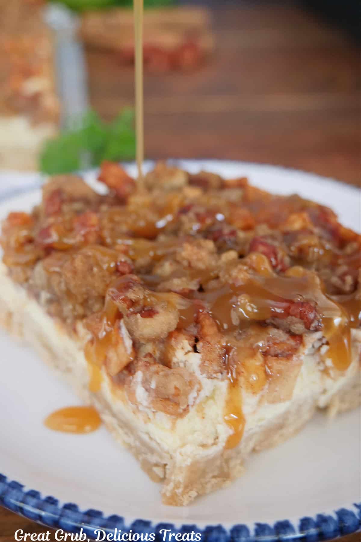 A round white plate with blue trim with a serving of a caramel apple cheesecake bar with caramel being drizzled on top.