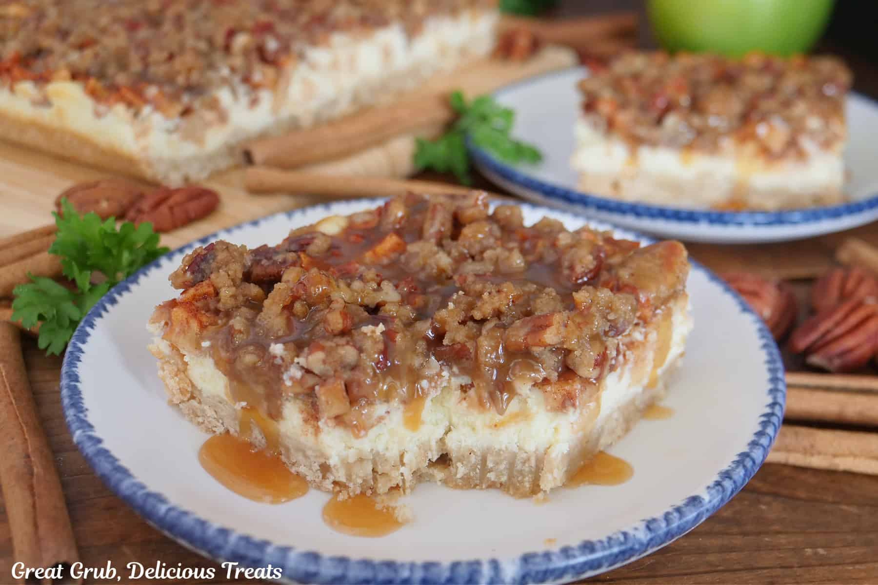 A round white plate with blue trim with a slice of apple cheesecake with caramel on top with a few bites taken out of it with another piece in the background.