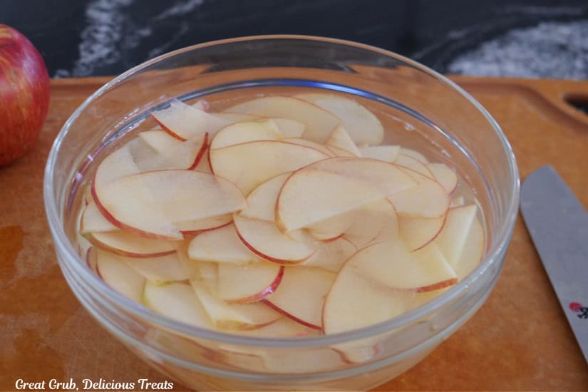 A wood surface with a glass bowl filled with thinly sliced apples soaking in lemon water.