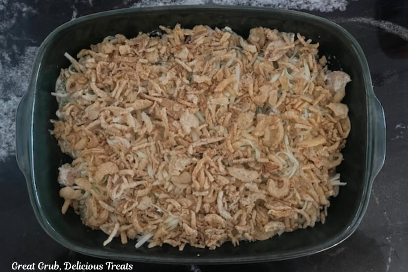 A dark green baking dish with chicken, green beans, cheese, a creamy sauce and topped with crispy fried onions.