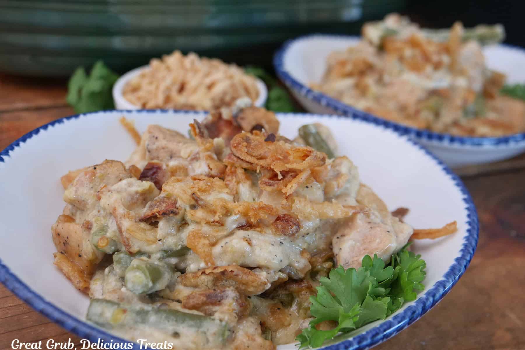 A wood surface with two white bowls with blue trim filled with a serving of chicken green bean casserole in them.