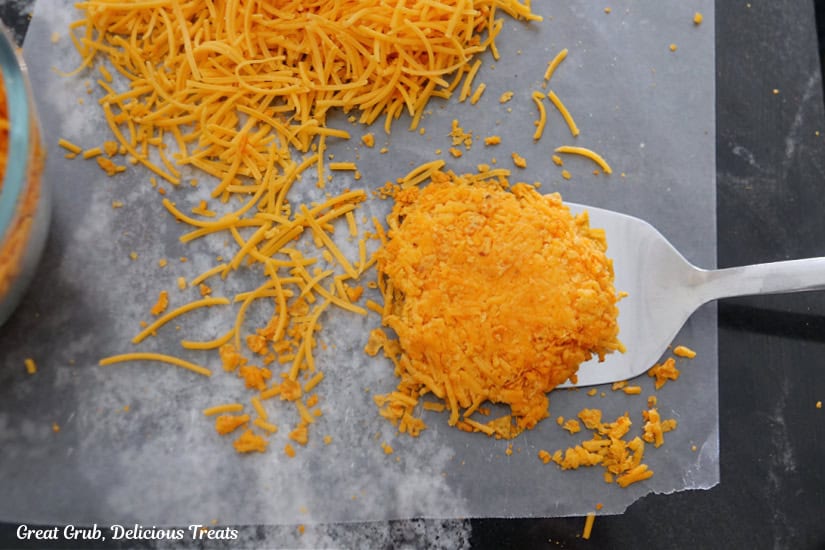 A black counter top with parchment paper on it with shredded cheese and crushed Nacho cheese Doritos on it made into a "tortilla."