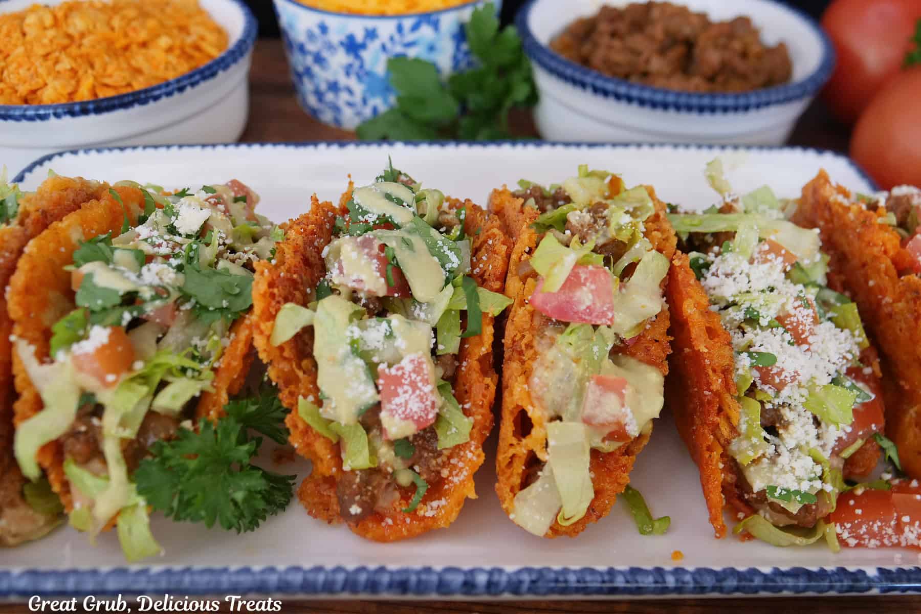 A rectangle white plate with blue trim with six mini cheesy crunch tacos on it.
