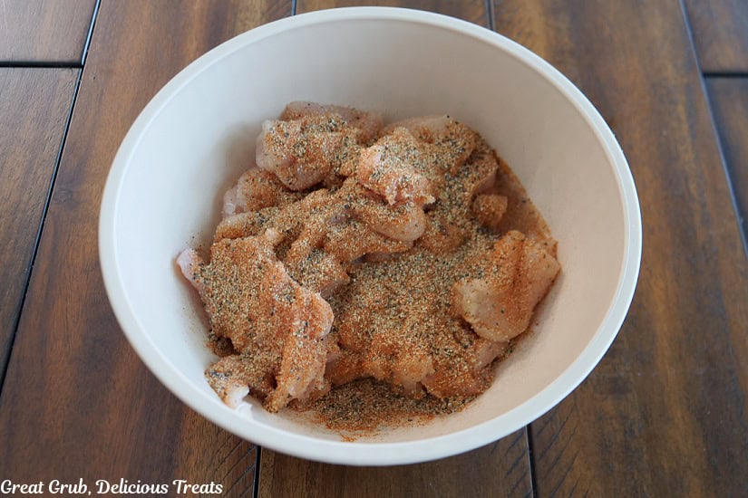 A wooden table with a white bowl filled with sliced chicken, onions, and seasonings in it.