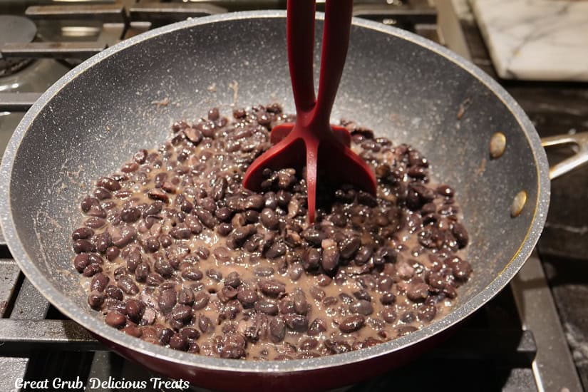 A frying pan with black beans in it and they are being mashed up a bit.