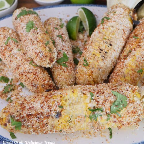 A white plate with blue trim with six grilled ears of corn with the ingredients for Mexican street corn on them.