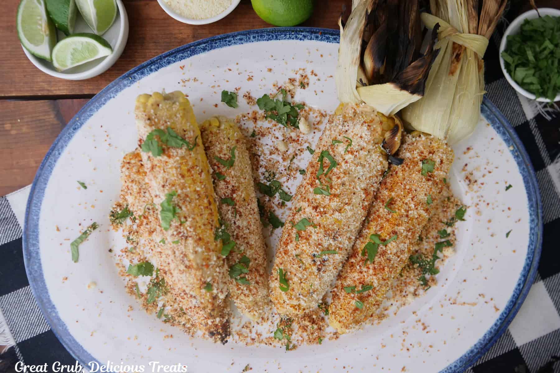 A wood surface with a white round plate with blue trim with six ears of grilled corn on the cob coated with Mexican street corn ingredients.