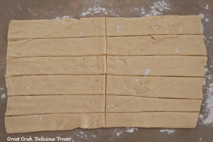 Parchment paper on a counter top with crescent dough in a rectangle that has been cut into 6 long strips, and then they are cut in half.