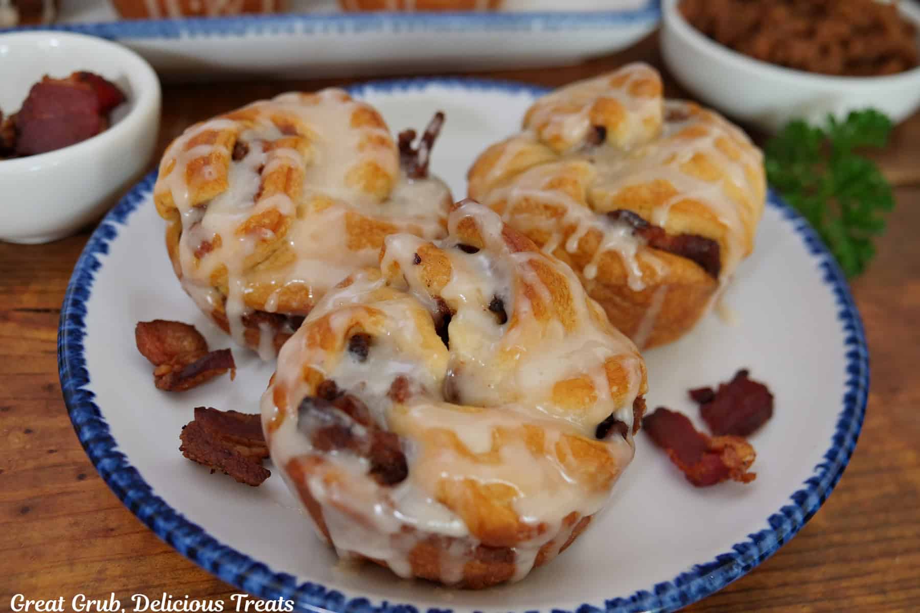A wood surface with a round white plate with blue trim with three maple bacon muffins on it.