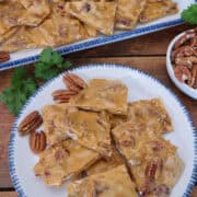 A white plate with blue trim with pieces of homemade pecan brittle on it with more in the background, and a title overlay at the top.