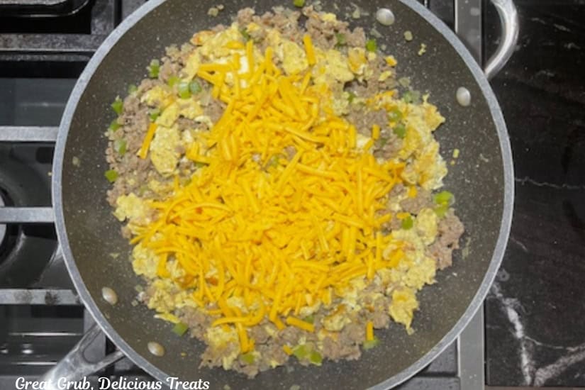 A frying pan with cooked scrambled eggs and sausage mixture with shredded sharp cheddar cheese on it.