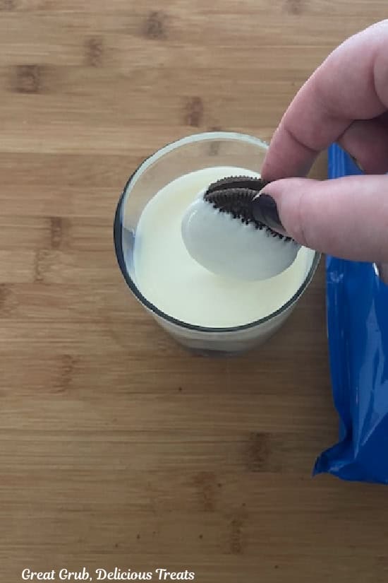 A wood surface with a cup filled with melted white chocolate in it with an Oreo being dipped into the chocolate.