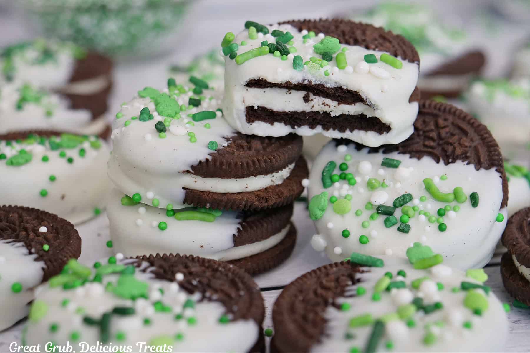 A white surface with white chocolate dipped Oreos with festive St Patrick's Day sprinkles on them and a bite taken out of the cookie on top.