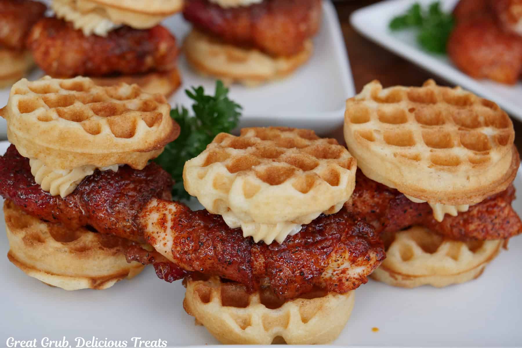 A white plate with three mini bacon wrapped chicken and waffles on it with more in the background.