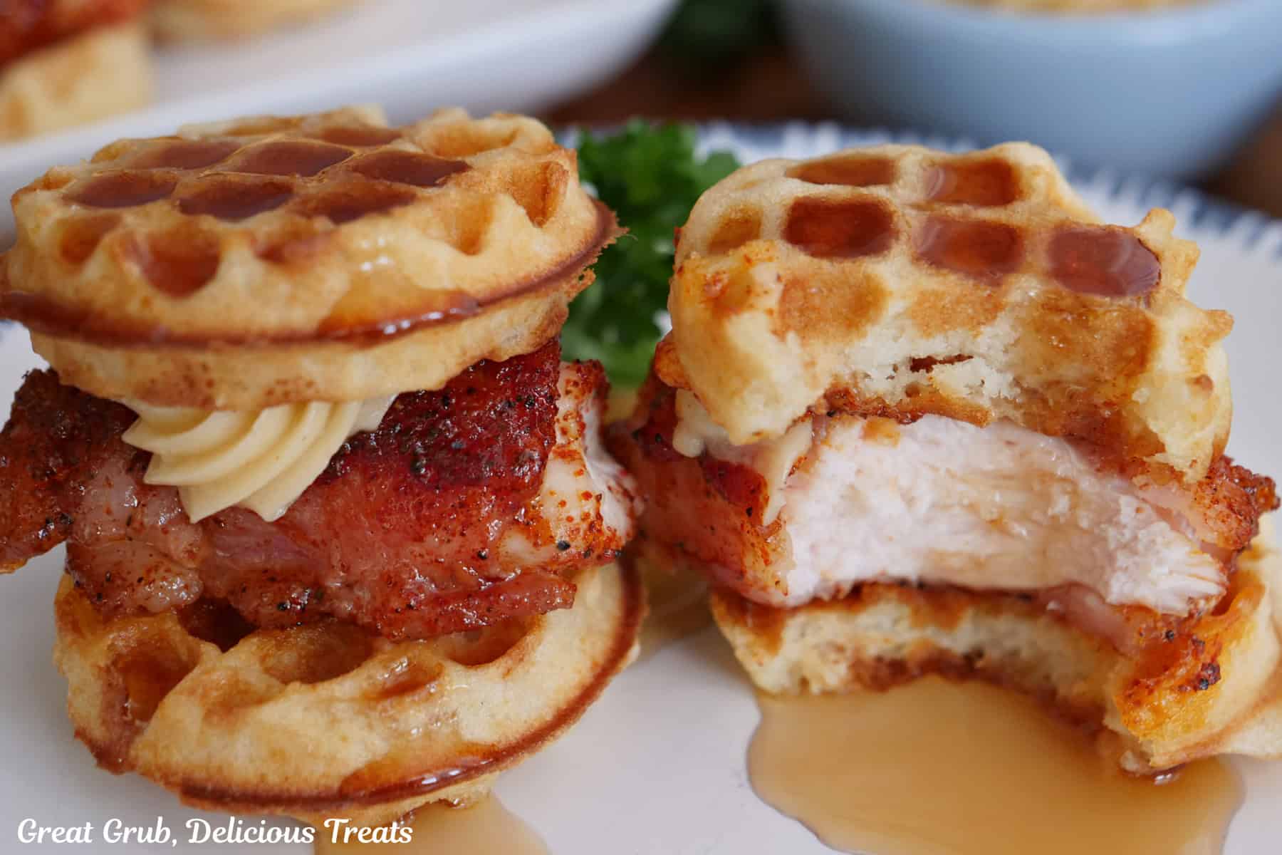 A white plate with two mini bacon wrapped chicken and waffles with honey butter with a bite out of one of the mini waffles.
