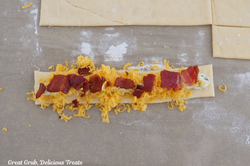 A strip of crescent dough with a layer of cream cheese mixture, shredded cheddar cheese, and crumbled, crispy bacon on top.