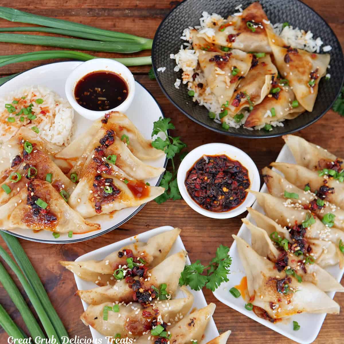 A wood surface with three white plates and a black bowl filled with chili crisp pork potstickers with two small white bowls filled with dipping sauce, and green onions place on the surface.
