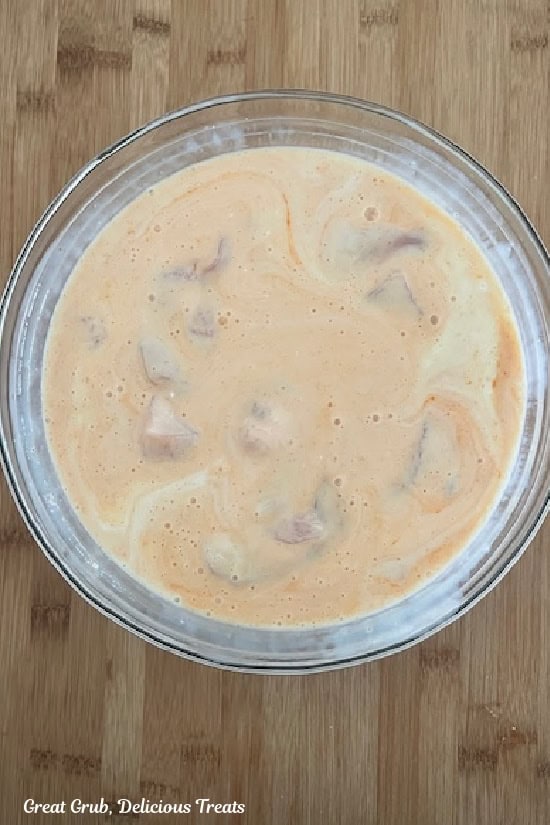 A wood surface with a glass bowl filled with buttermilk, pickle juice, hot sauce, and chicken pieces that are marinating.