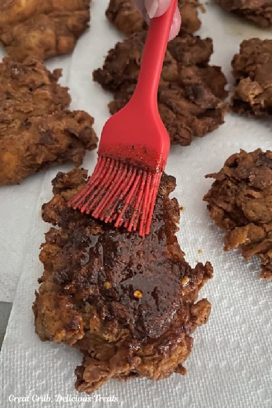 Paper towels on a surface with fried pieces of chicken on them and a hot seasoned oil being brushed over the tops and bottom of each piece of chicken.