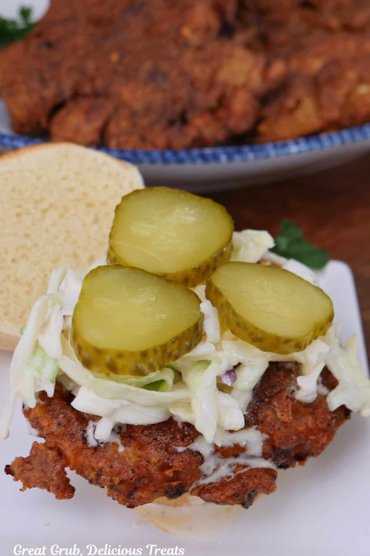 A white small plate with a slider bun opened up with a fried piece of chicken on it with topped with coleslaw and three sliced pickles.