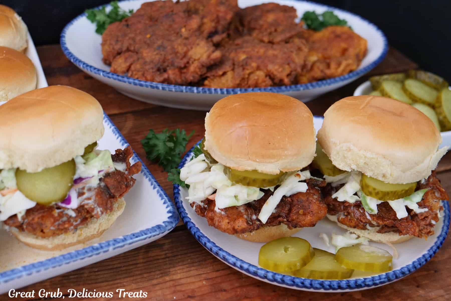 A wood surface with two white plates with blue trim with Nashville hot chicken sliders on them and another oval white plate with blue trim with fried chicken pieces on it.