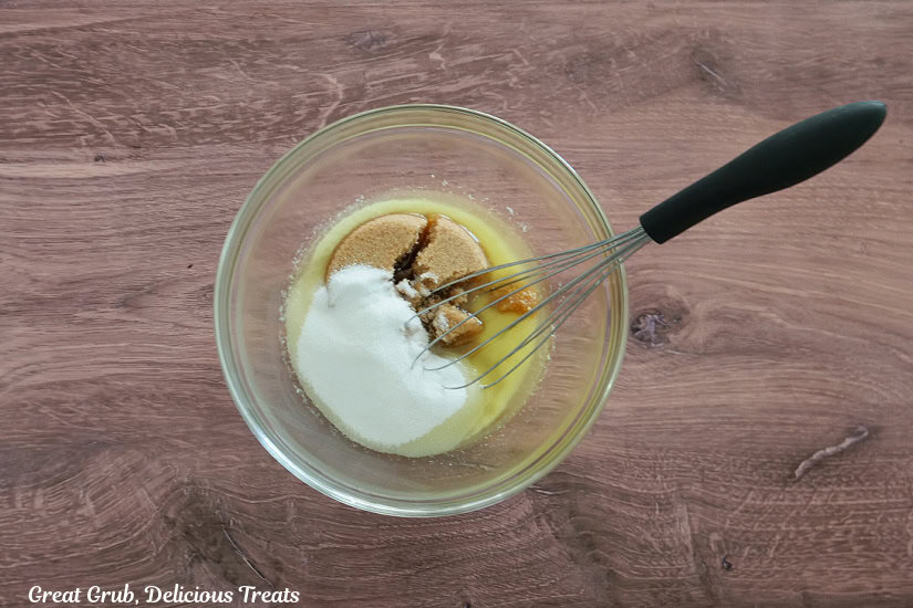 A wood surface with a glass bowl filled with melted butter and sugars, and a whisk leaning up on the edge of the bowl before the mixture is being mixed together.