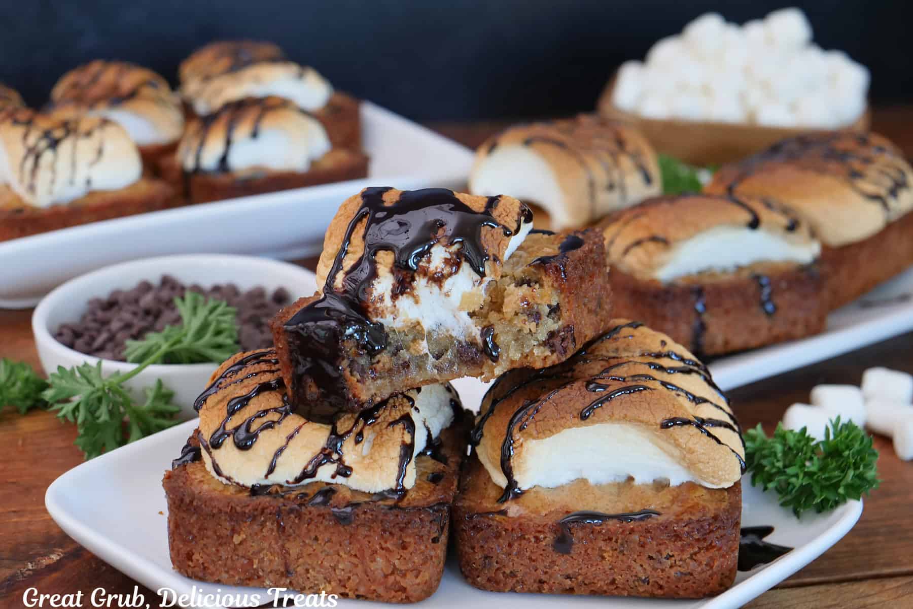 A wood surface with S'mores blondies on white plates with chocolate drizzled of each blondie.