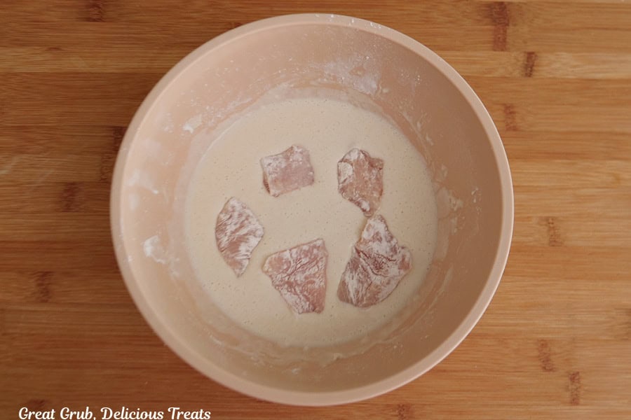 A wood surface with a light pink bowl with chicken pieces being soaked into the wet mixture.