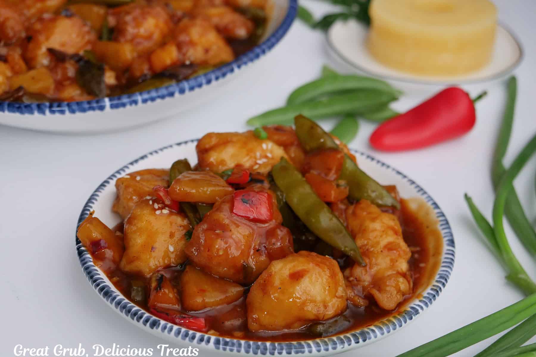 A white surface with a white bowl with blue trim filled with a serving of chicken stir-fry in it with another bowl in the background, and green beans, a red pepper, and pineapple rings in the background.