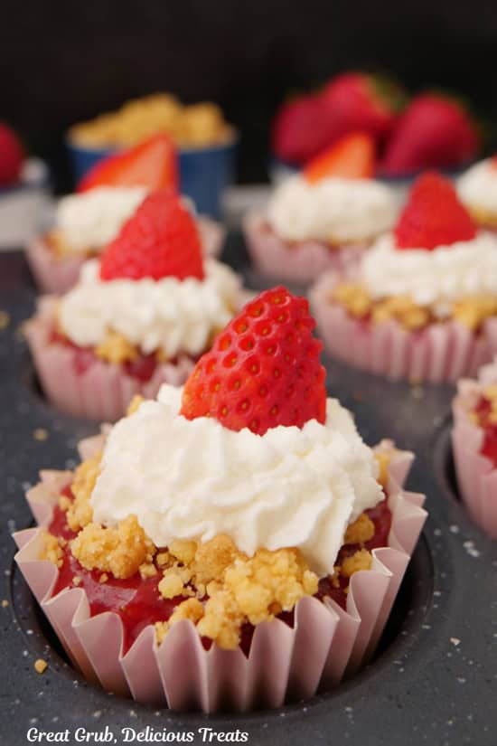 A muffin tin with the mini strawberry lemon cheesecakes in it topped with whipped cream and a slices of strawberries.