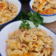 A wood surface with three white bowls with blue trim filled with bacon mac and cheese, and a title overlay at the top.