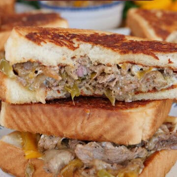 A white plate with blue trim with a cheesesteak grilled cheese sandwich on it that is cut in half and a bite taken out of the slice on top, and a title overlay at the top.