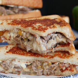 A white plate with blue trim with two halves of a cheesesteak grilled cheese on it with a bite taken out of the top half.