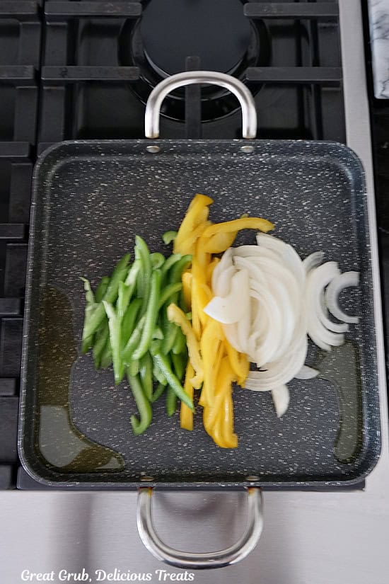 A grill pan with green and yellow bell pepper strips and onion strips on it before being grilled.