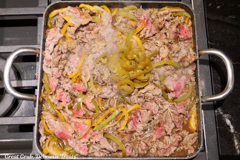 A grill pan with the onions, peppers, shaved beef, and jalapeno slices added to the pan.