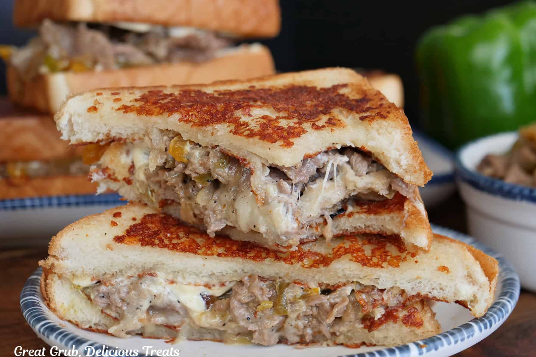 A round white plate with blue trim with two halves of grilled cheesesteak sandwiches with a bite taken out of the piece on top.